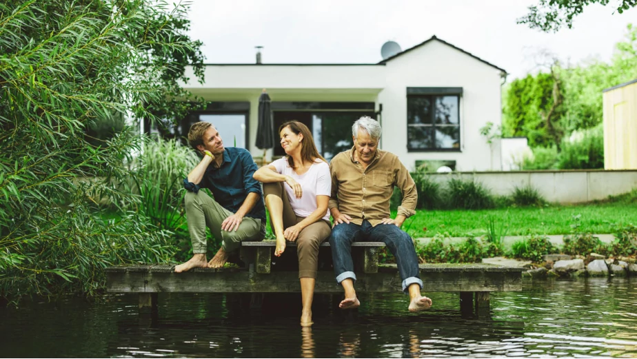 Familia de tres adultos sentados al aire libre junto a un arroyo que sonríen y disfrutan de un día soleado.