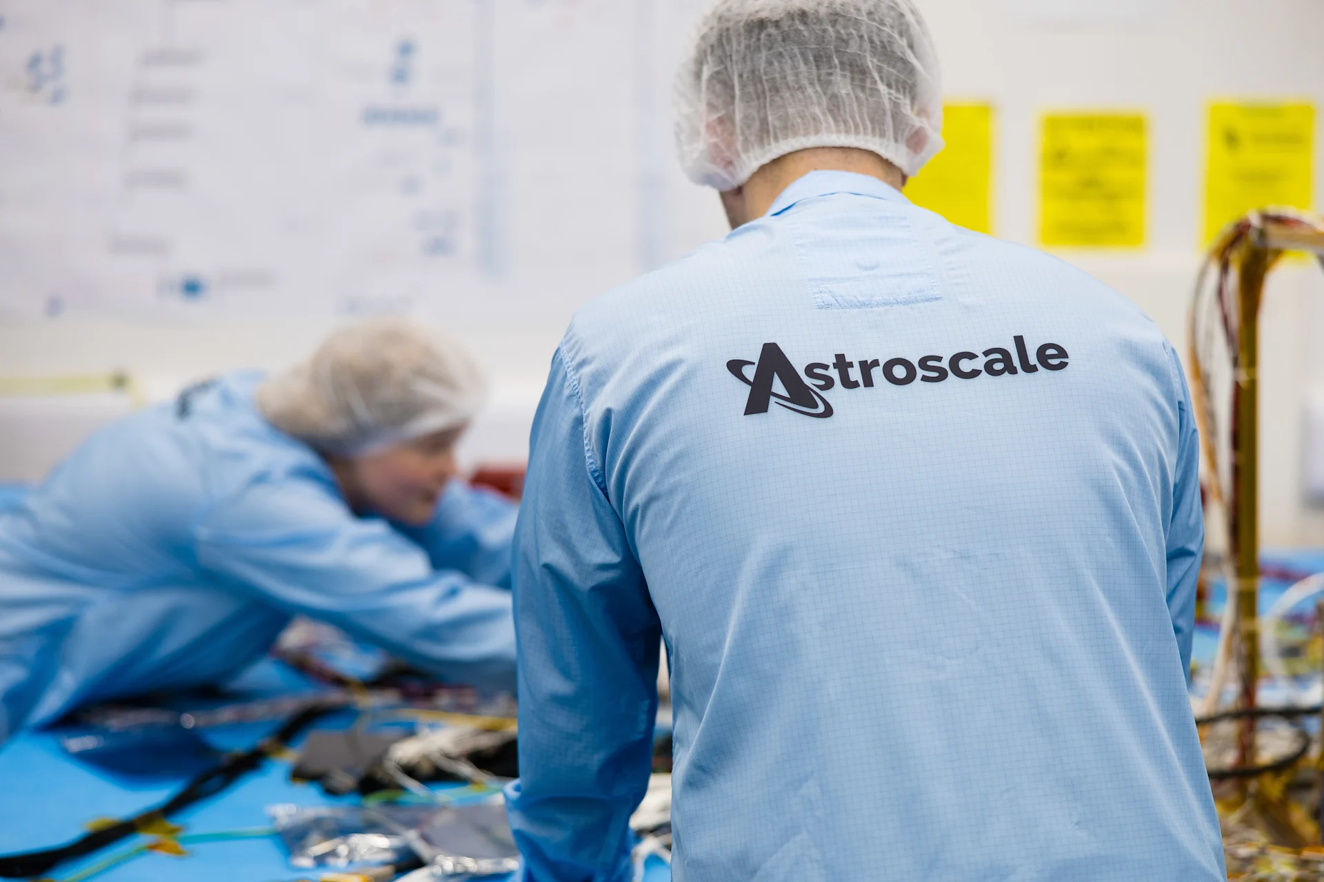 Two Astroscale engineers in blue cleanroom suits and hair nets working on satellite components in a cleanroom