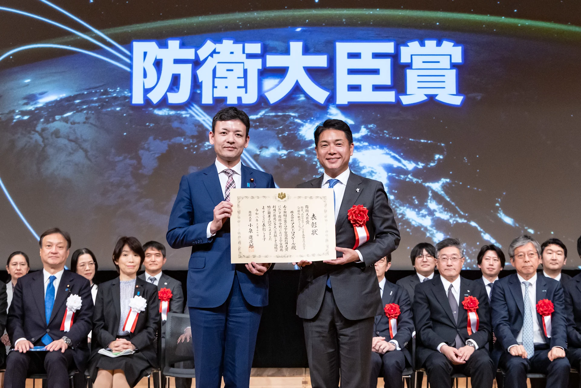 日本の防衛式典において、アストロスケールCEO兼創業者である岡田光信が防衛大臣賞の賞状を受け取っている様子。背景には関係者が着席している。
