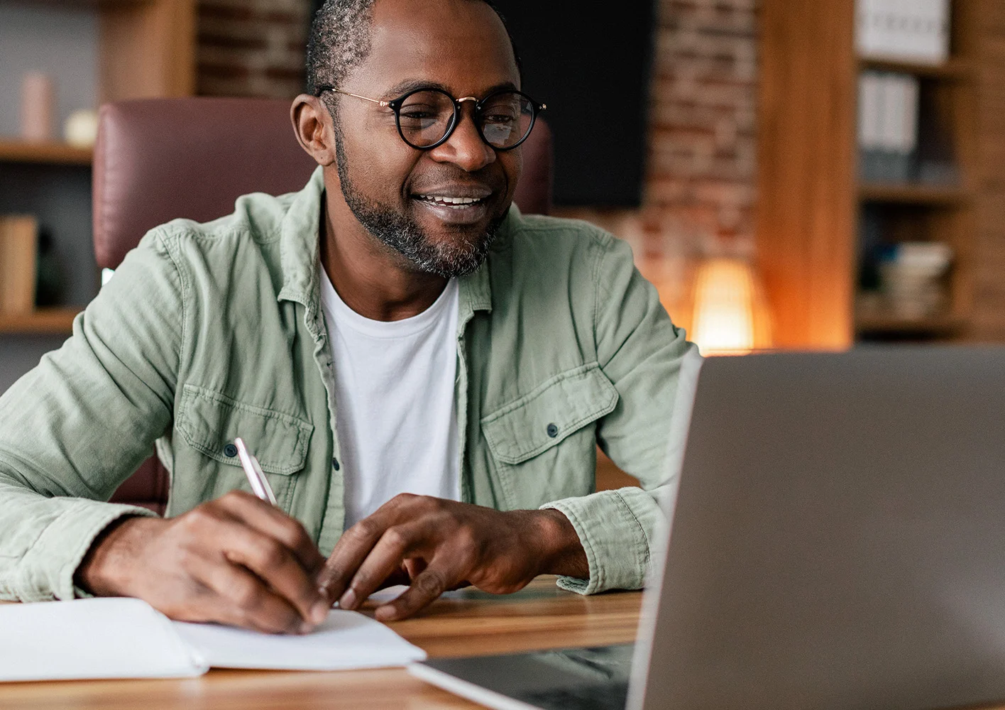 Man sitting at desk, looking at laptop and writing in a notebook