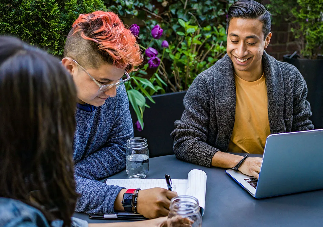 Three colleagues seated outside at a table with a bench, surrounded by green plants and purple flowers.