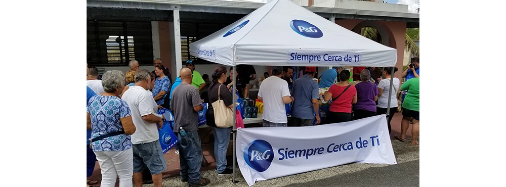 一眾市民在印有 P&G 標誌及「Siempre Cerca de Ti」（常伴左右） 字樣的白色帳篷下排隊，等候領取由 P&G 及其合作夥伴提供的賑災物資與服務。