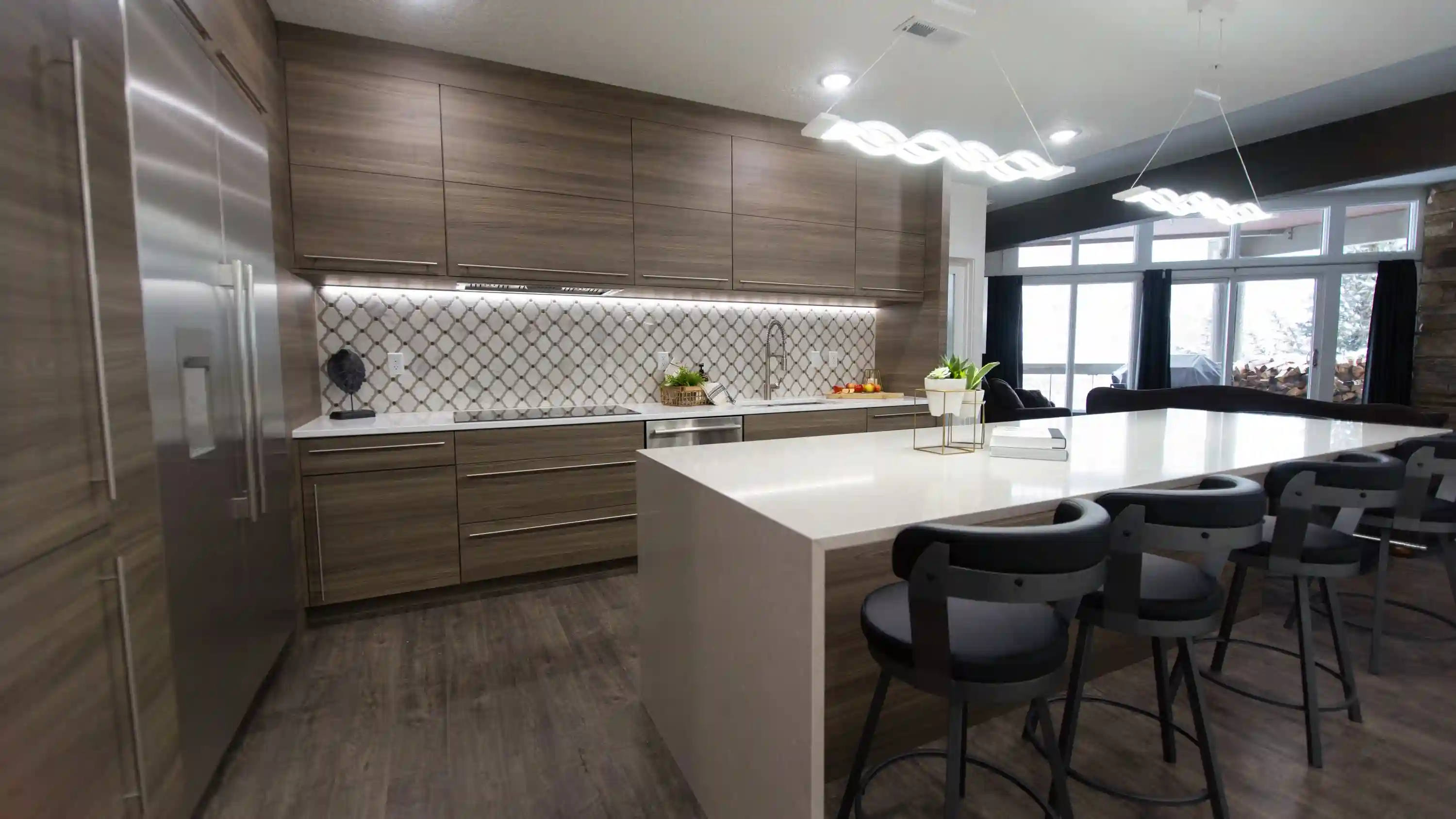 A newly remodeled kitchen. A mosaic tile back splash is lit up by lights lining under the cabinetry. White countertops contrast against mixed wood toned cabinetry. A kitchen isladn with a quartz top stands out. Two statement lights hang over the island in a wavy pattern.