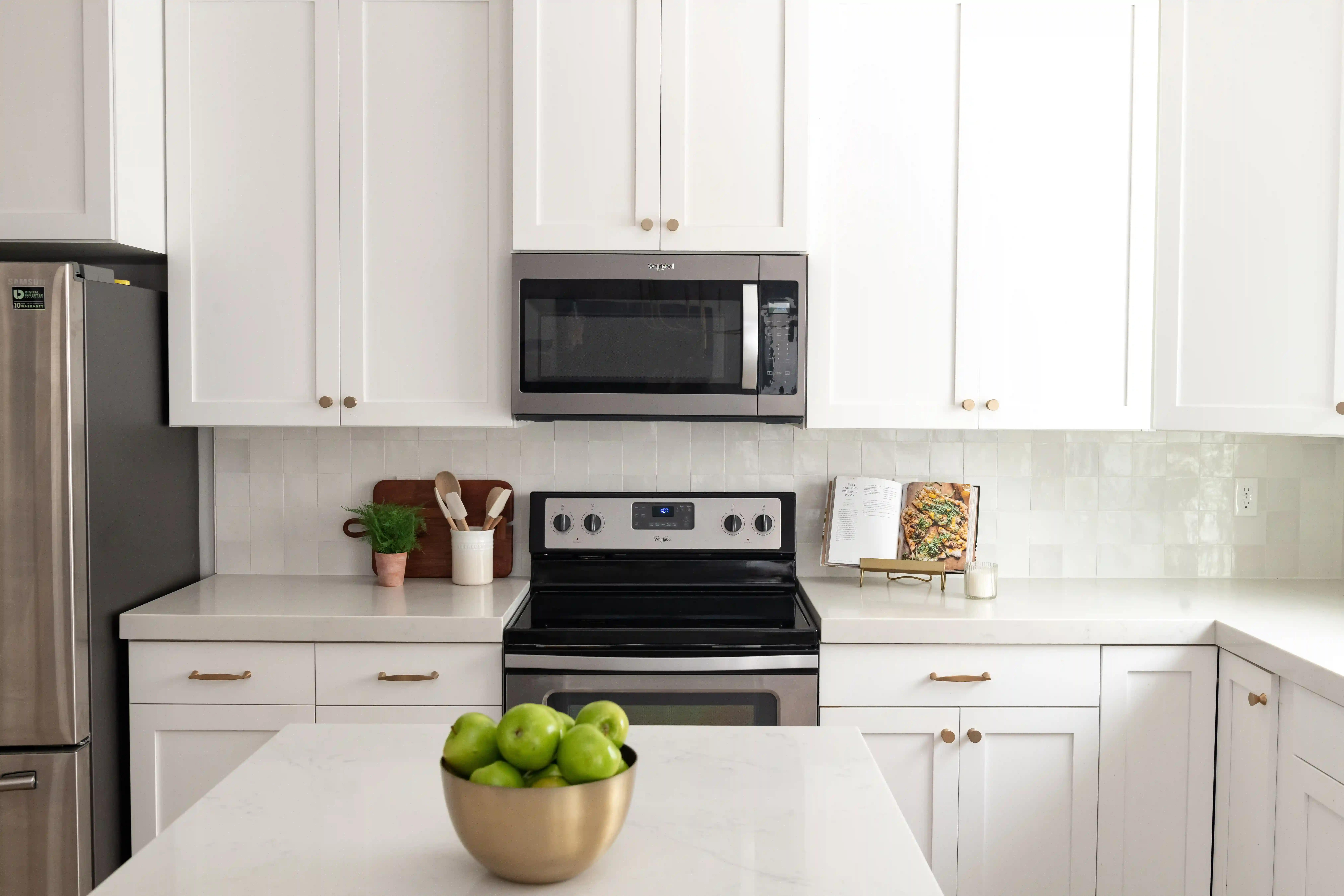 A kitchen with a creamy textured white backsplash between white upper and lower cabinets with brass handles. White countertops contrast with the stainless steel oven and microwave in the center of the wall. 
