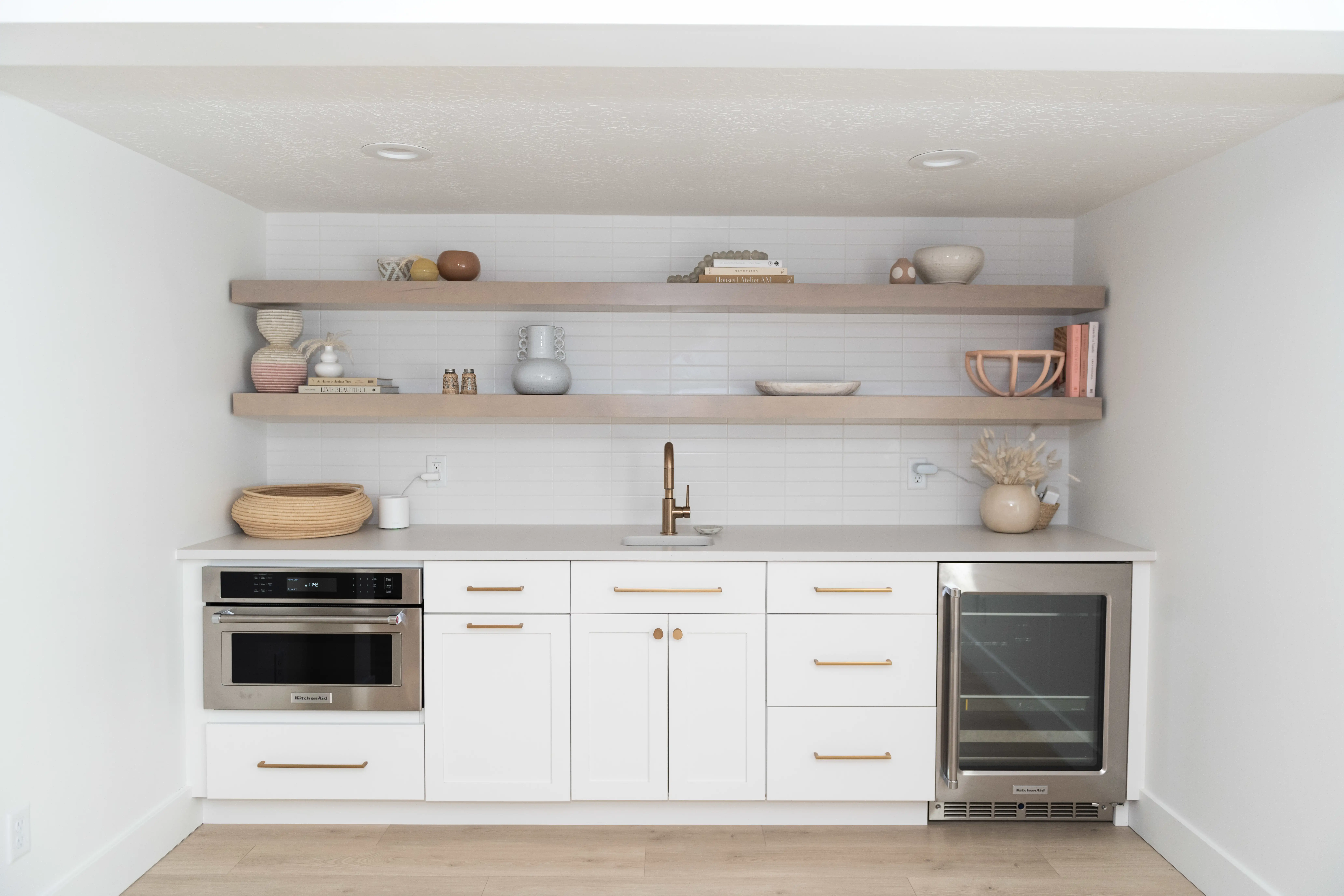 A kitchenette with two full length wood shelves over the countertop. White subway tile backsplash makes the shelves pop. The kitchenette has white counters and cabinets with brass handles and hardware. A warm wood flooring spans the whole room.
