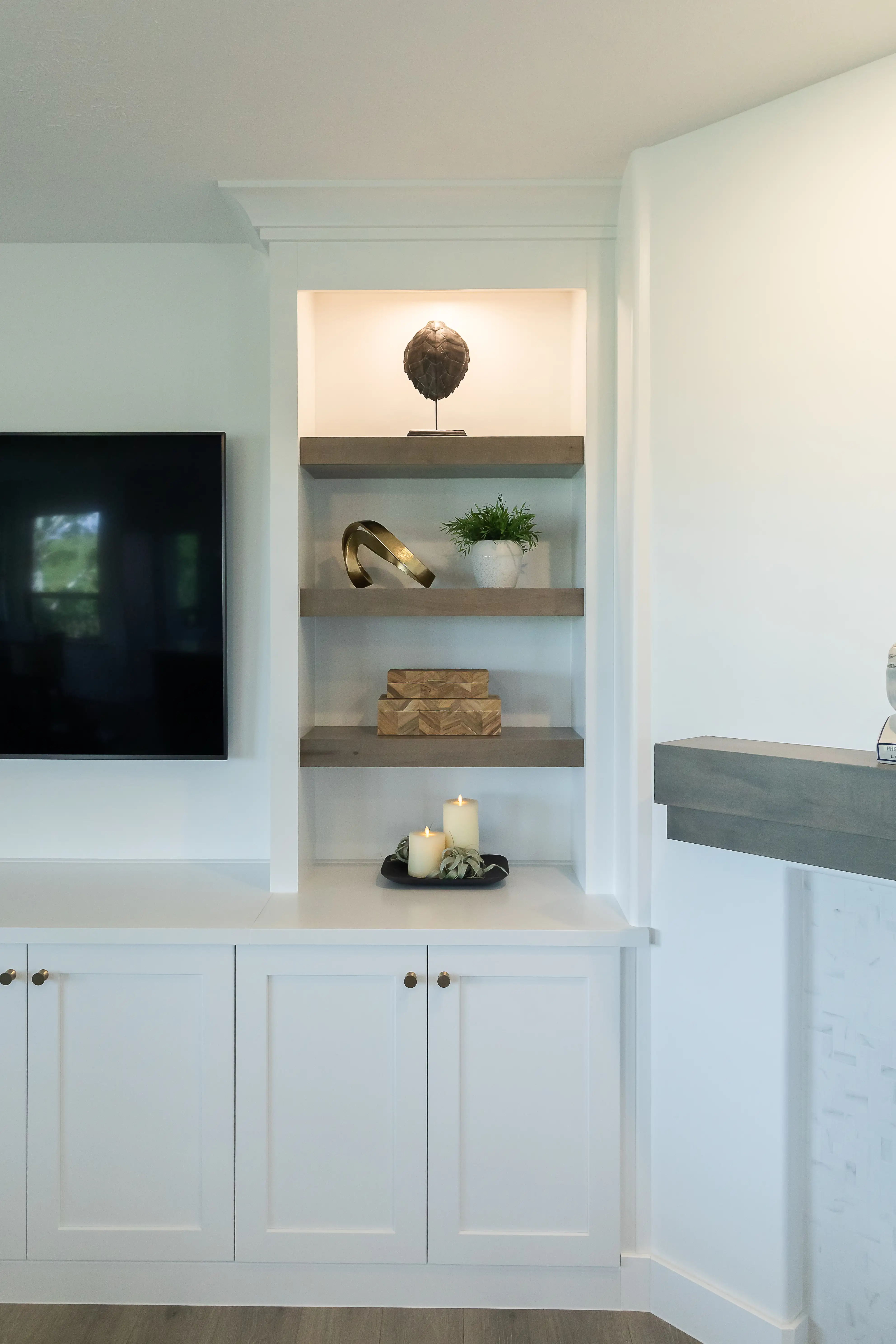 Three wooden shelves are installed into a wall beside a wall mounted TV. Lighting is integrated into the nook and decorative objects are placed upon each shelf. 