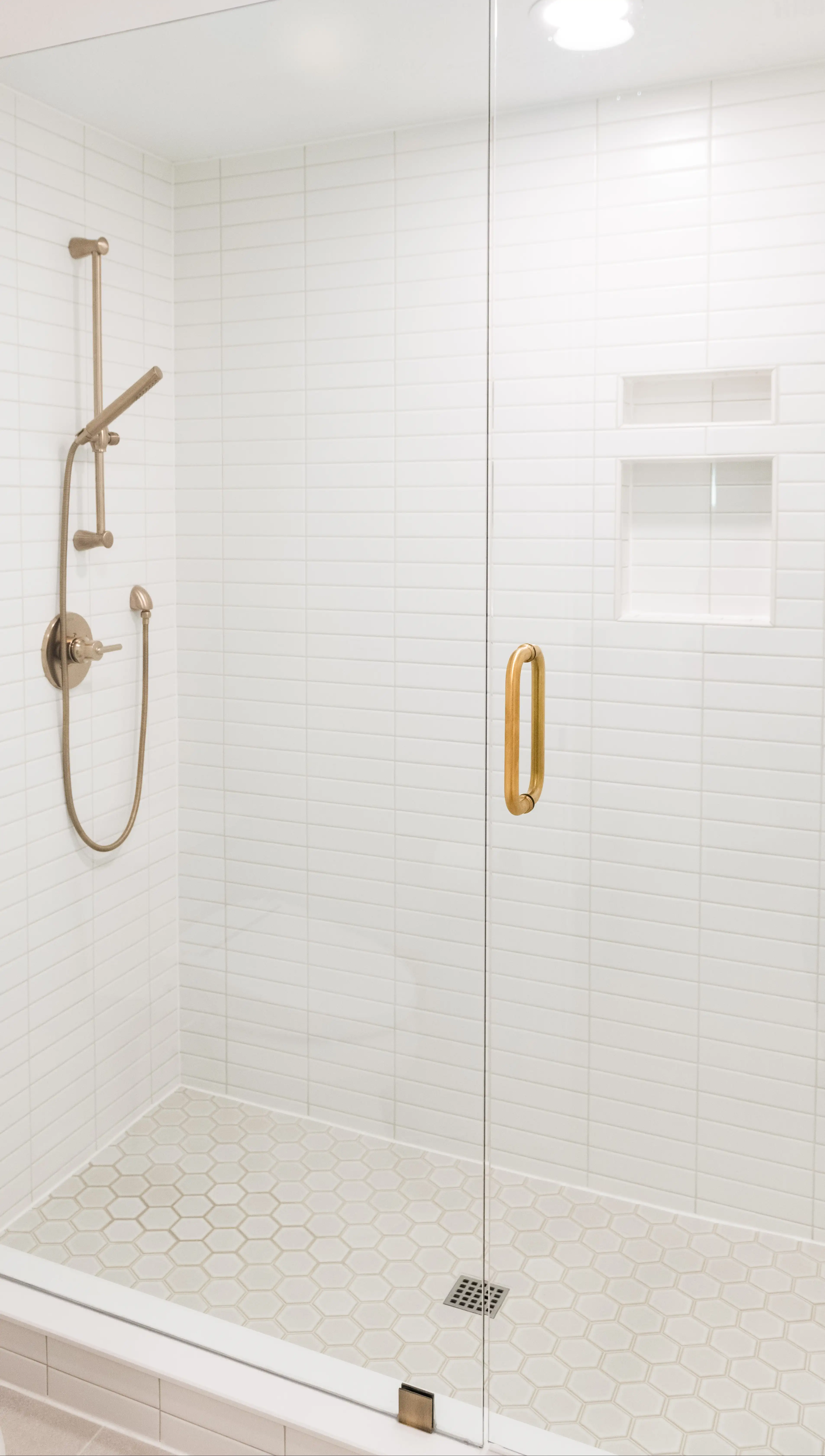 A white tile shower with a cream hexagon tile floor, a clear glass panel and door with brass fixtures. 