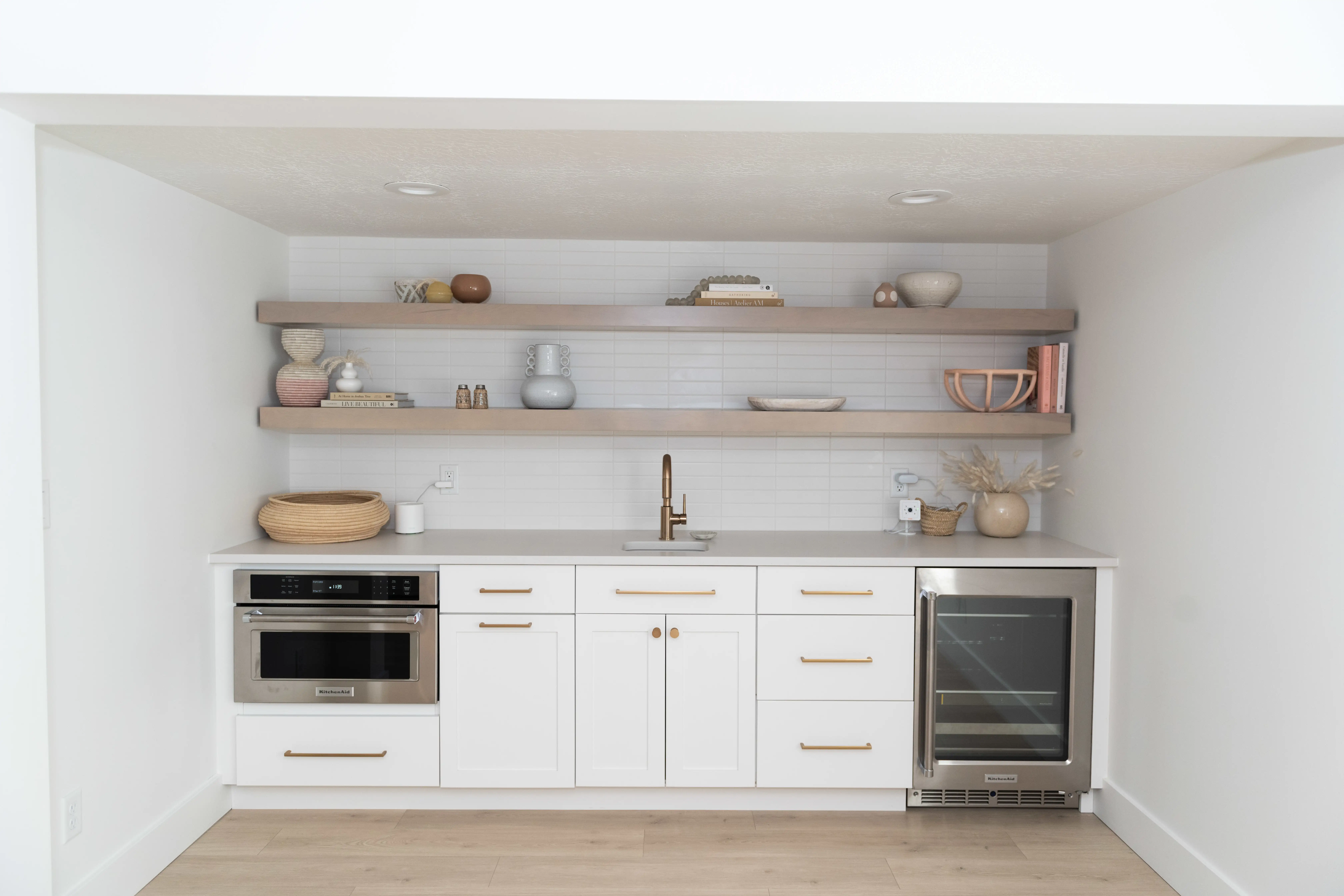 Two full length wooden shelves hang above a kitchenette that spans from wall to wall. White cabinetry and countertops with brass hardware and handles. The flooring is a neutral, warm toned wood like vinyl flooring.