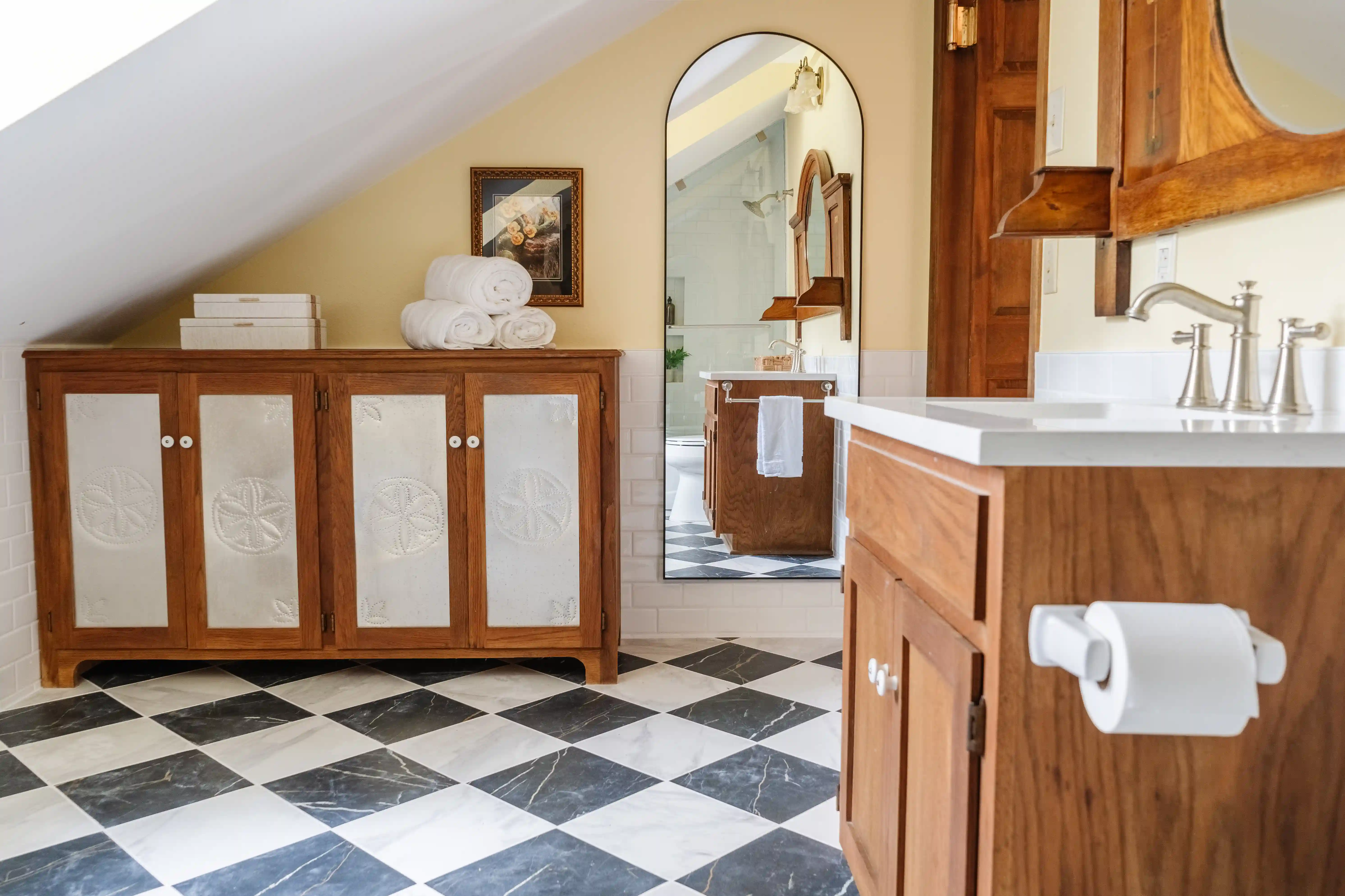 A bathroom with marble black and white checkered floors, a tan back wall, and a white slanted ceiling. There are two two door natural wood cabinets with white panels that have sand dollars engraved into them and silver handles to each door. Placed on the top of them is a stack of white books on the left and three towels stacked into a triangle on the right of the top of the cabinets. Behind the three towels on the wall is a wooden framed picture on the wall that is undecernable as to what it is in the photo. To the right of the cabinet and picture is a full length bosy mirror with an arched top and square bottom corners. You can see a wooden door that closely matches the two cabinets on the left of the wall. Coming closer to the audience looking at the photo on the right wall of the room is a wooden framed mirrow over a bathroom vanity with a white counter top, brushed satin faucet with two handles and wooden cabinets with two brushed satin handles on each cabinet door. 