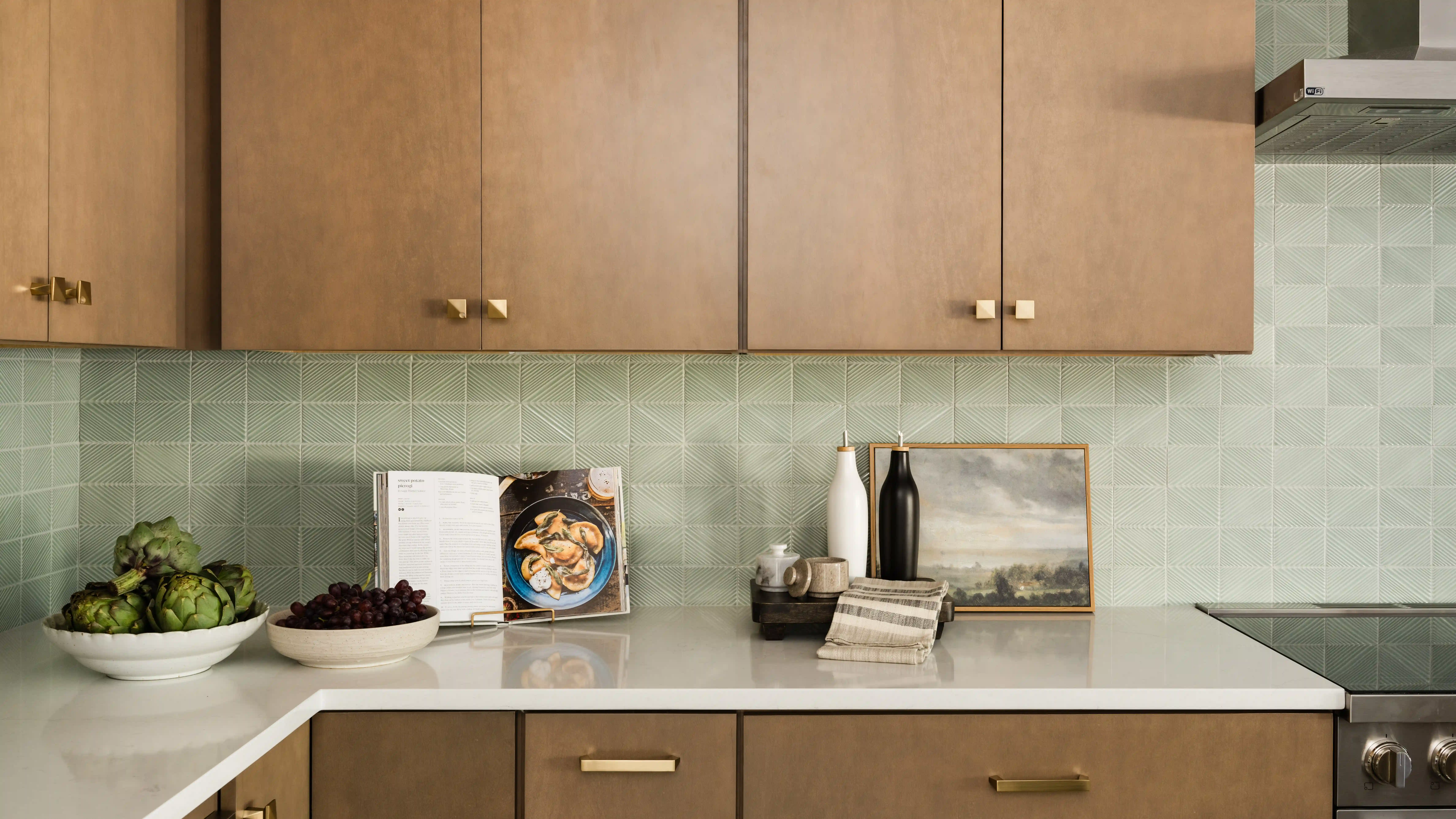 Natural wooden toned upper and lower cabinets pop against a sage green square tile backsplash and white countertops. 