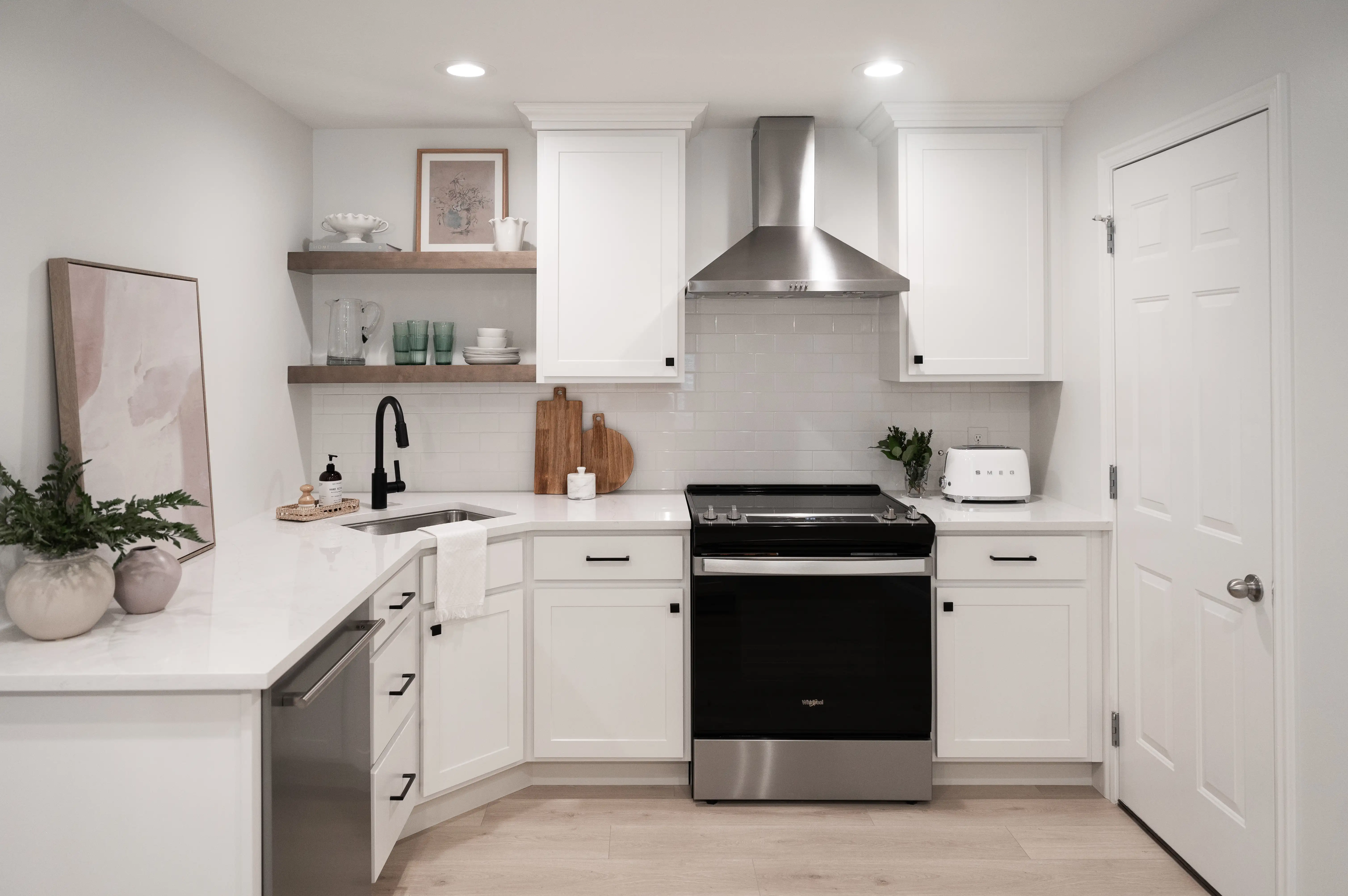 A kitchenette with white cabinets, counters, and subway tile backsplash. Stainless steel appliances throughout. A light neutral finish wood like vinyl flooring warms up the space. A black faucet and hardware pull everything together in the kitchenette. 