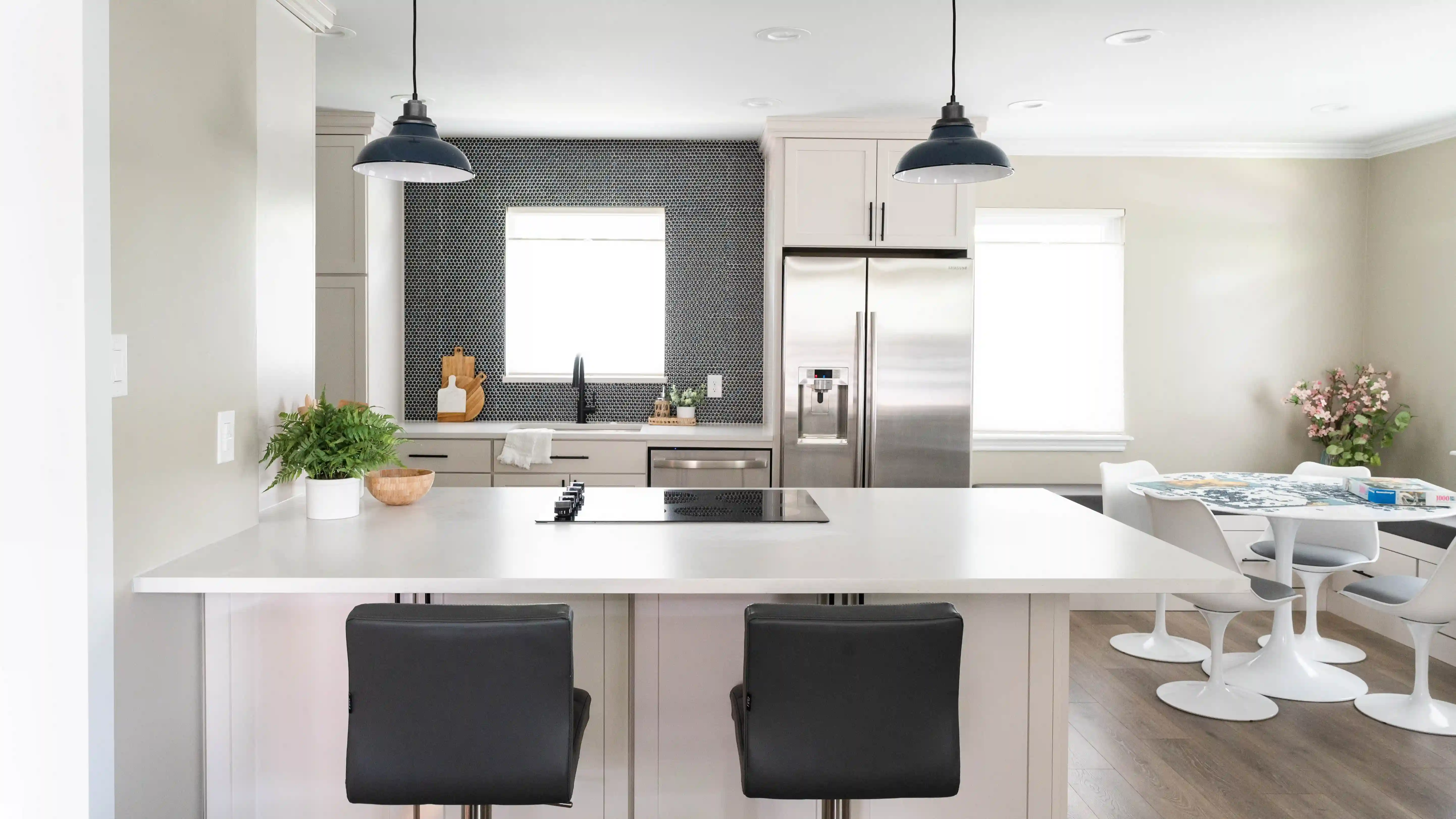 Two black leather barstools sit up against a white kitchen island with white countertops. A white table and chairs sits in the right side corner of the kitchen. All the appliances within the kitchen are stainless steel. A black penny tile backsplash contrasts the white of the kitchen on the back wall over the sink. 