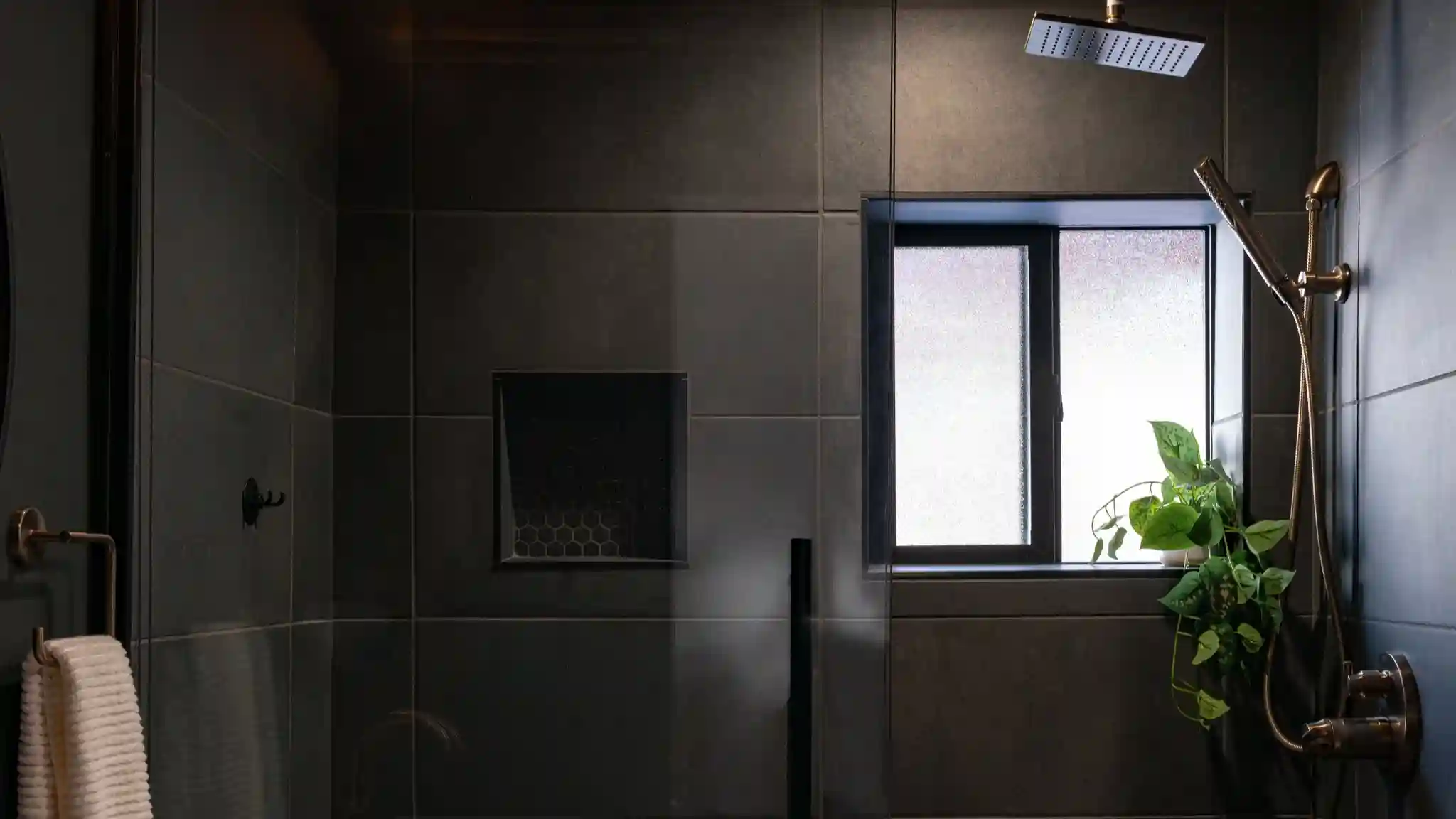 An all black tiled shower with a slliding glass door. The hardware is brass and contrasts against all of the black. A small window lets in some natural light into the bathroom.