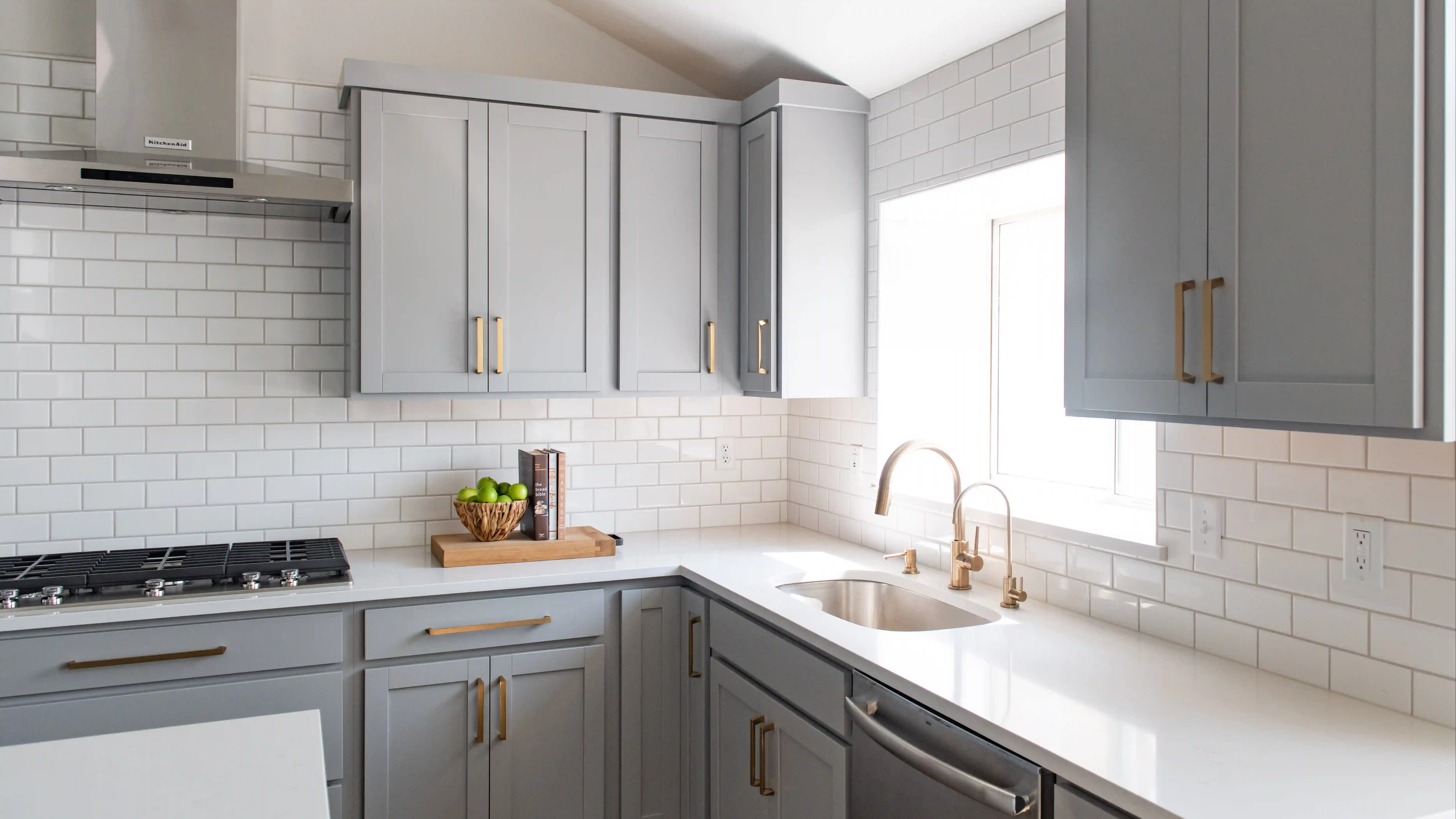 A kitchen with a white subway tile backsplash, white quartz countertops, and medium grey cabinetry with brass pulls. 