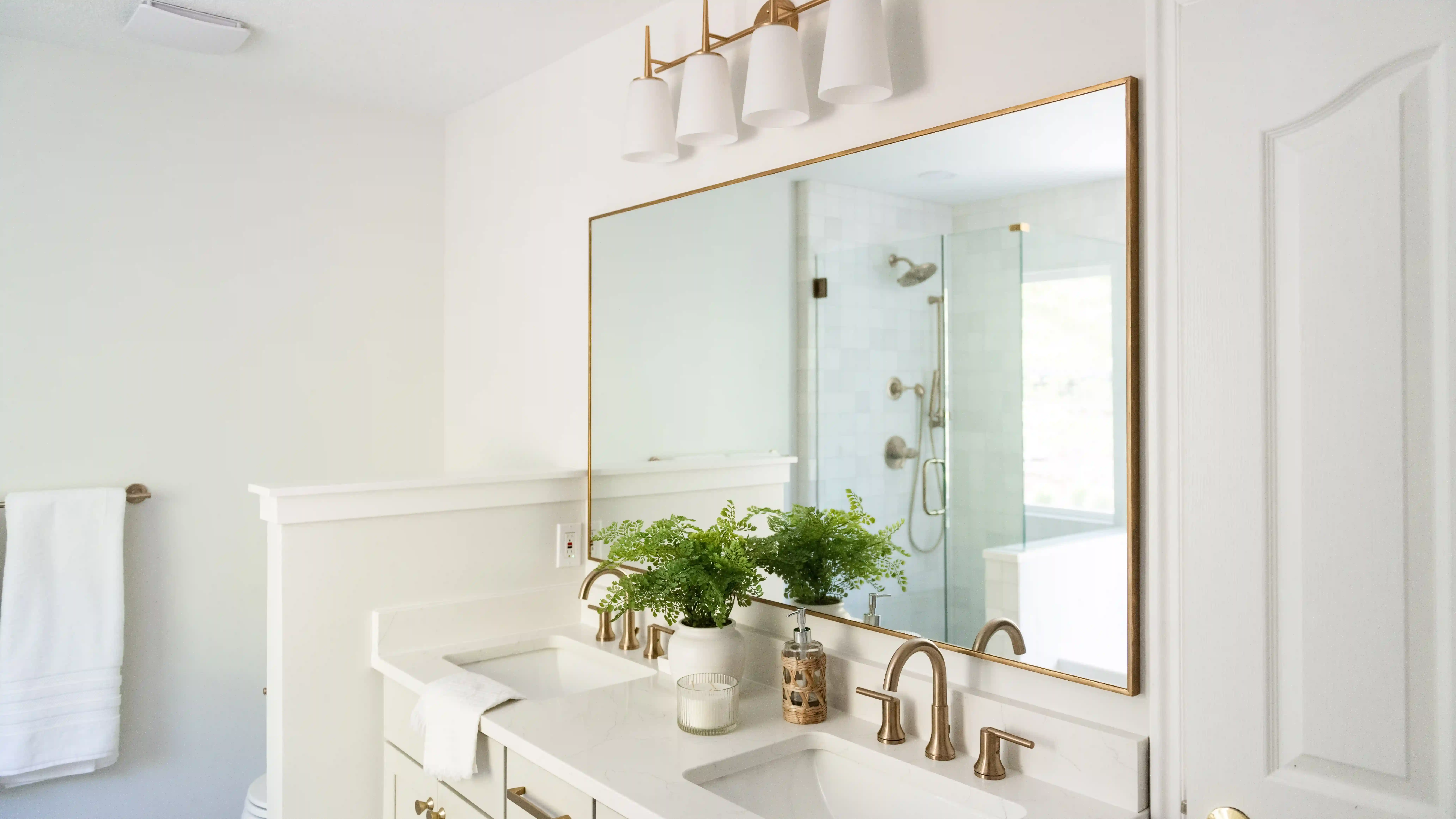 A bathroom with white walls, white countertops and double vanity, and brass accents in the faucets, mirror frame, and lighting. A wicker woven soap holder sits on the counter by each sink. A fern in a white vase with a candle sitting next to it is on the counter between sinks. 