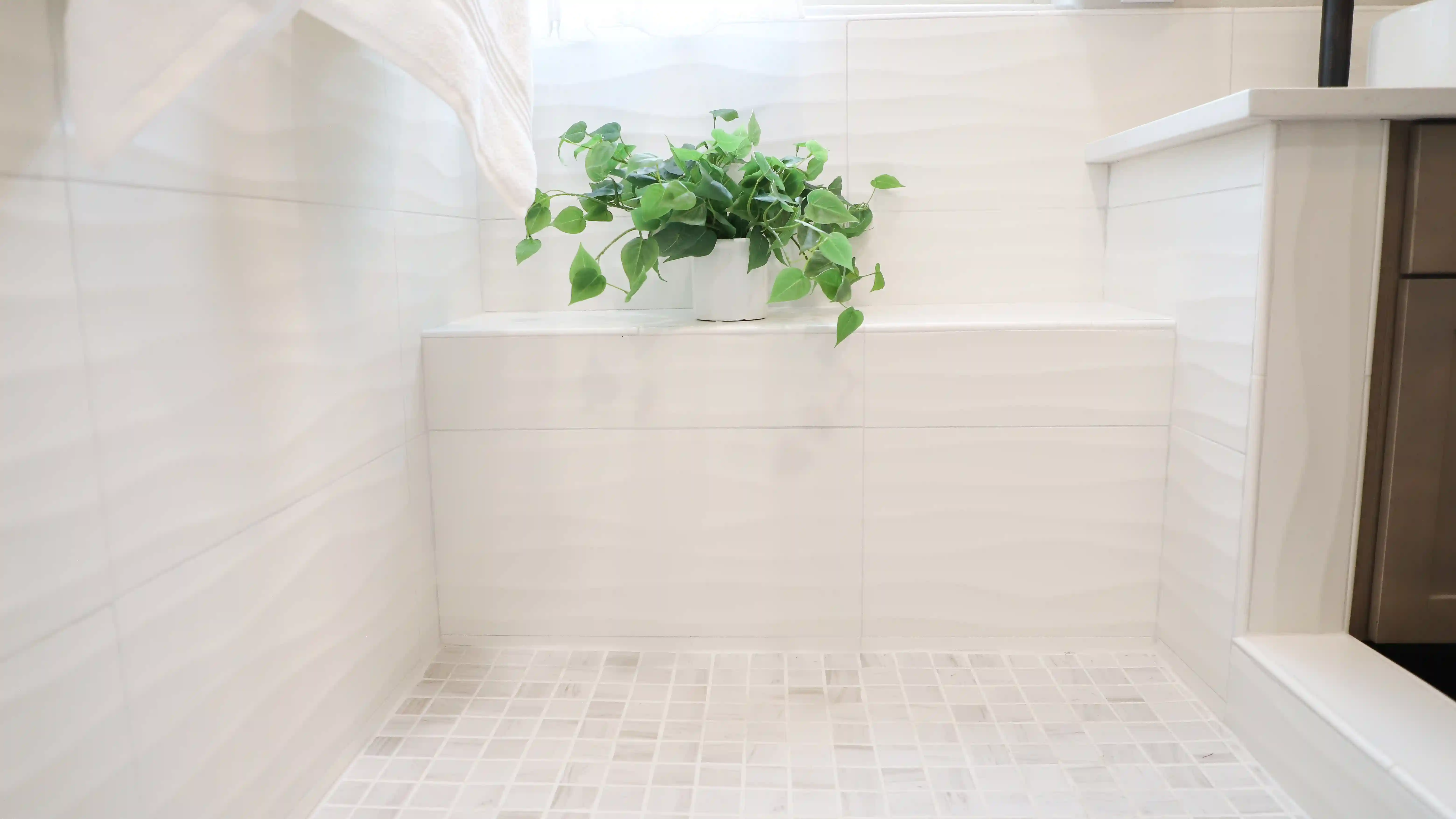 A shower with a shower pan tiled in neutral square tiles. The shower surround tiled with bog, wavy textured cream colored tiles. There is also a built in bench seat with matching textured tile. A house plant in a white vase sits on the left side of the bench in the shower. A window above the bench, lets in natural ight into the shower. 
