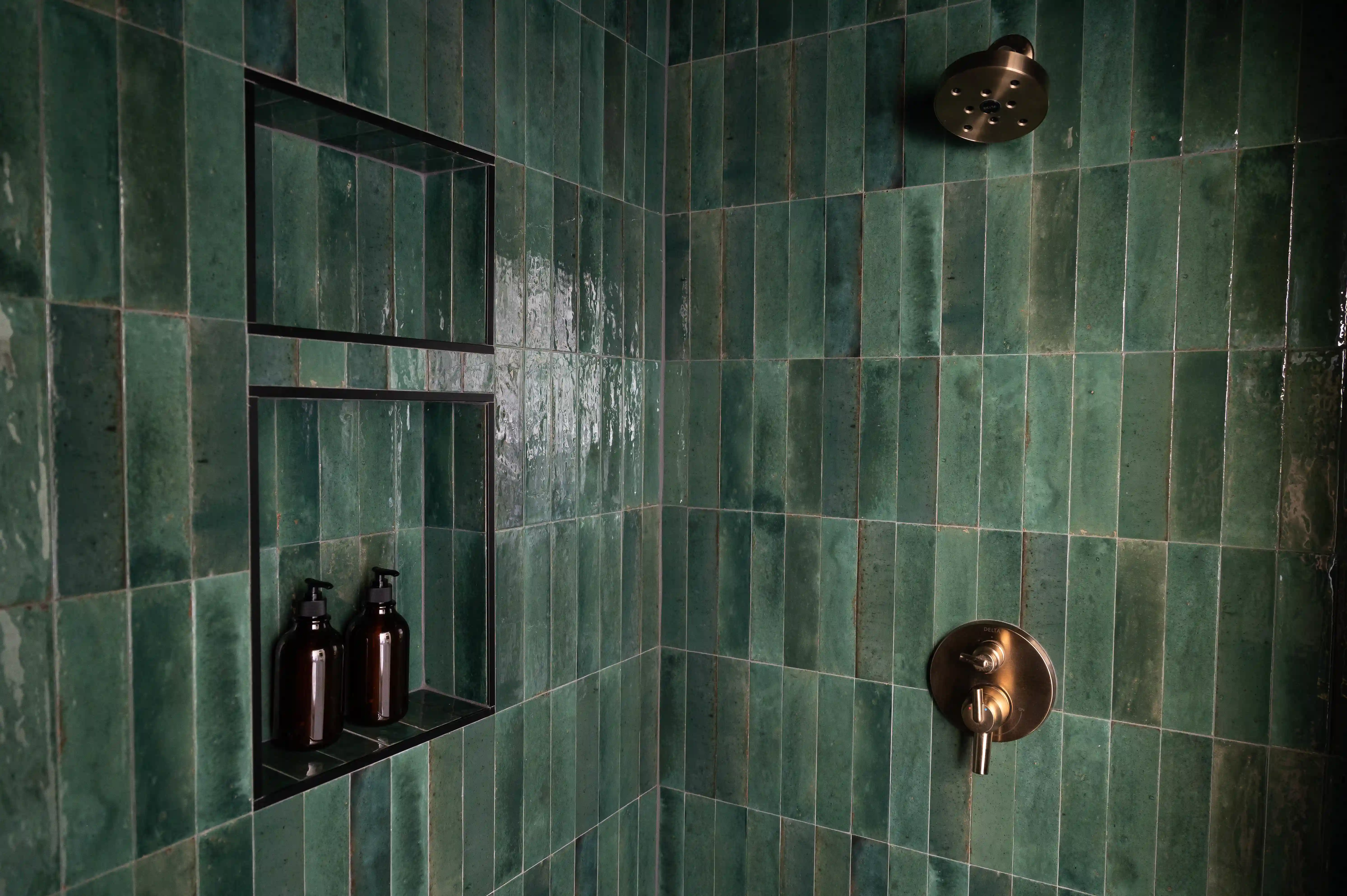 The Corner of a green tiled shower, top to bottom. The shower has two shelves built into the left wall with two amber glass bottles sitting on the bottom shelf. On the right side wall of the shower is a brass colored circular shower head with a matching handle below. 