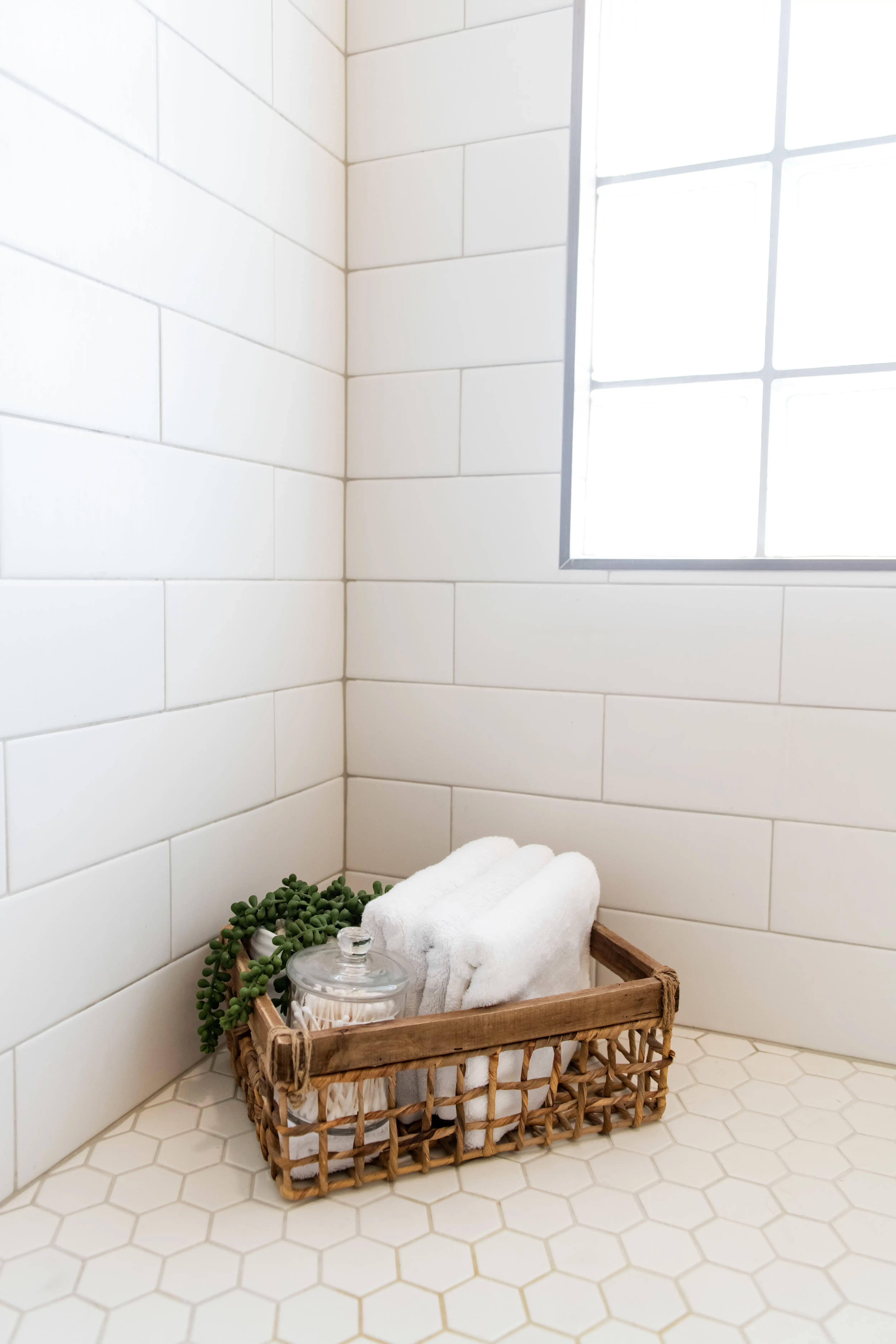 A corner in a bathroom with white subway tiled walls and white hex tiled flooring. A woven basket sits on the floor holding clean folded white towels, a succulent plant, and a glass canister. 