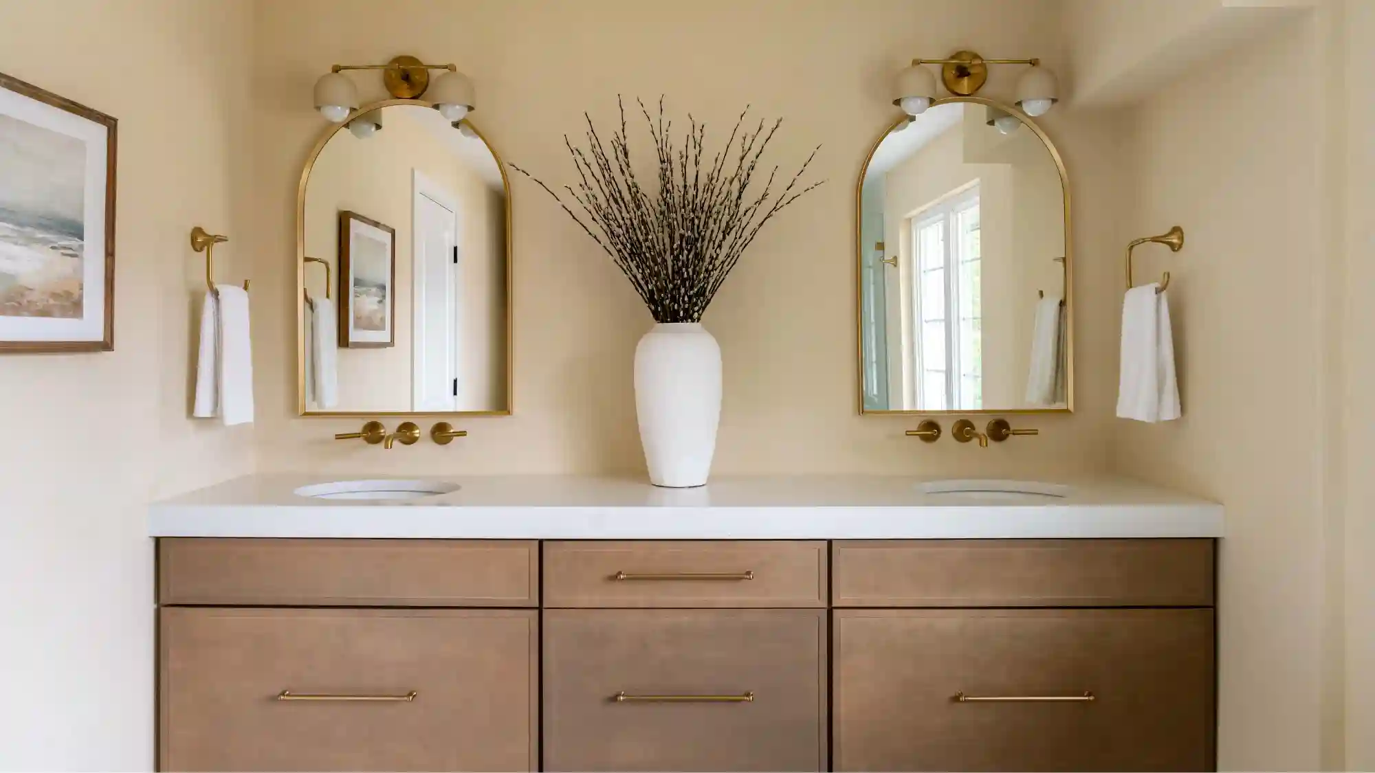 Vanity with two arched mirrors, decorative lighting, a large white vase with green stems. A neutral color palette and a light wash wood vanity with gold pulls. 