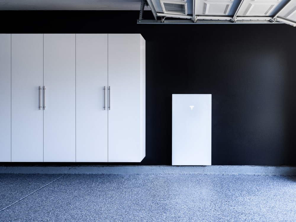 A clean and modern garage with the door fully open. On the black wall of the garage is a Tesla Powerwall 3 battery - a large white rectangle with a illuminated T Tesla logo in the top center.