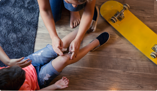 Adult applying a bandage to a child's scraped knee, with a skateboard lying nearby.