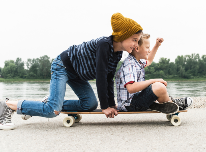 Two children enjoying a ride on a skateboard by a riverside, highlighting playful and active moments.