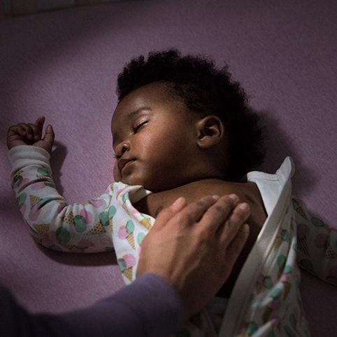 A sleeping baby in a colorful onesie lies on a pink surface, while an adult hand gently rests on the baby's chest.