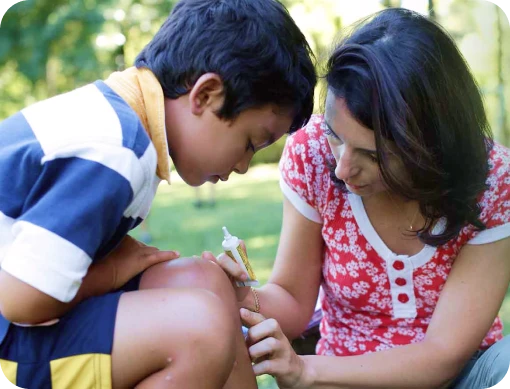 A mother applying NEOSPORIN to her son's knee while sitting outdoors, emphasizing the importance of wound care to prevent infections.