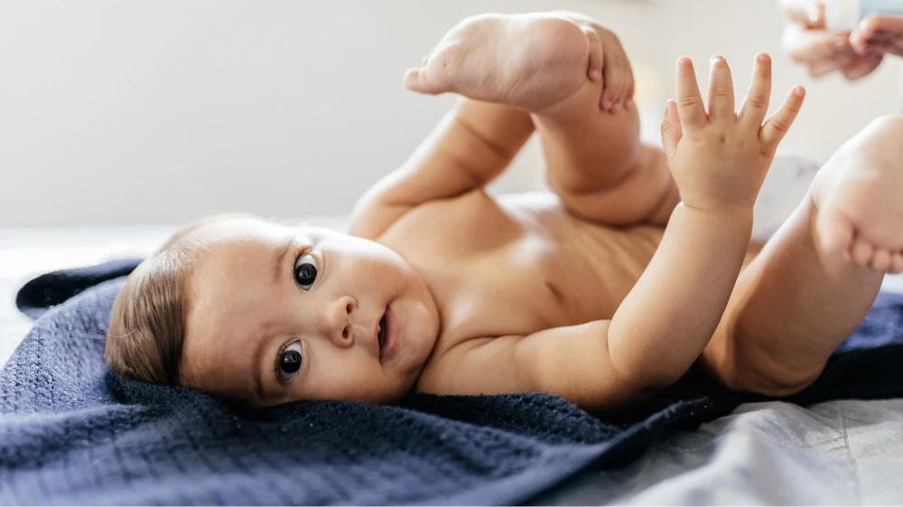 A baby lying on a dark blue blanket, with hands raised and feet in the air, playing and enjoying a moment of joy. Soft natural light illuminates the scene, creating a warm and inviting atmosphere.