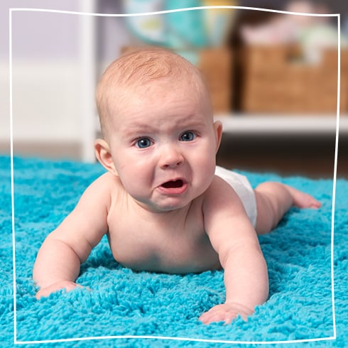 A baby in a diaper lying on a bright blue rug, looking up with a concerned or upset facial expression.