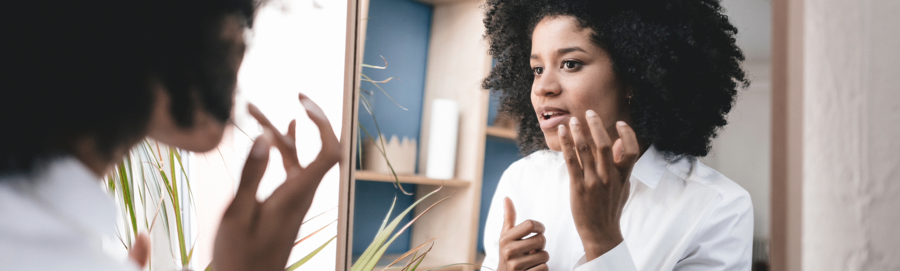 A woman with curly hair looking in a mirror and applying lip balm to treat chapped lips.