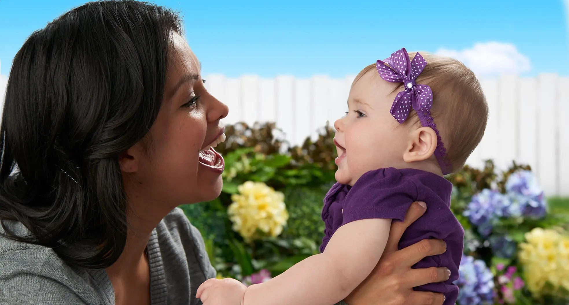 A mother and baby, both smiling, interact outdoors. The baby, dressed in purple with a bow headband, is held up by the mother against a background of flowers and a white fence.