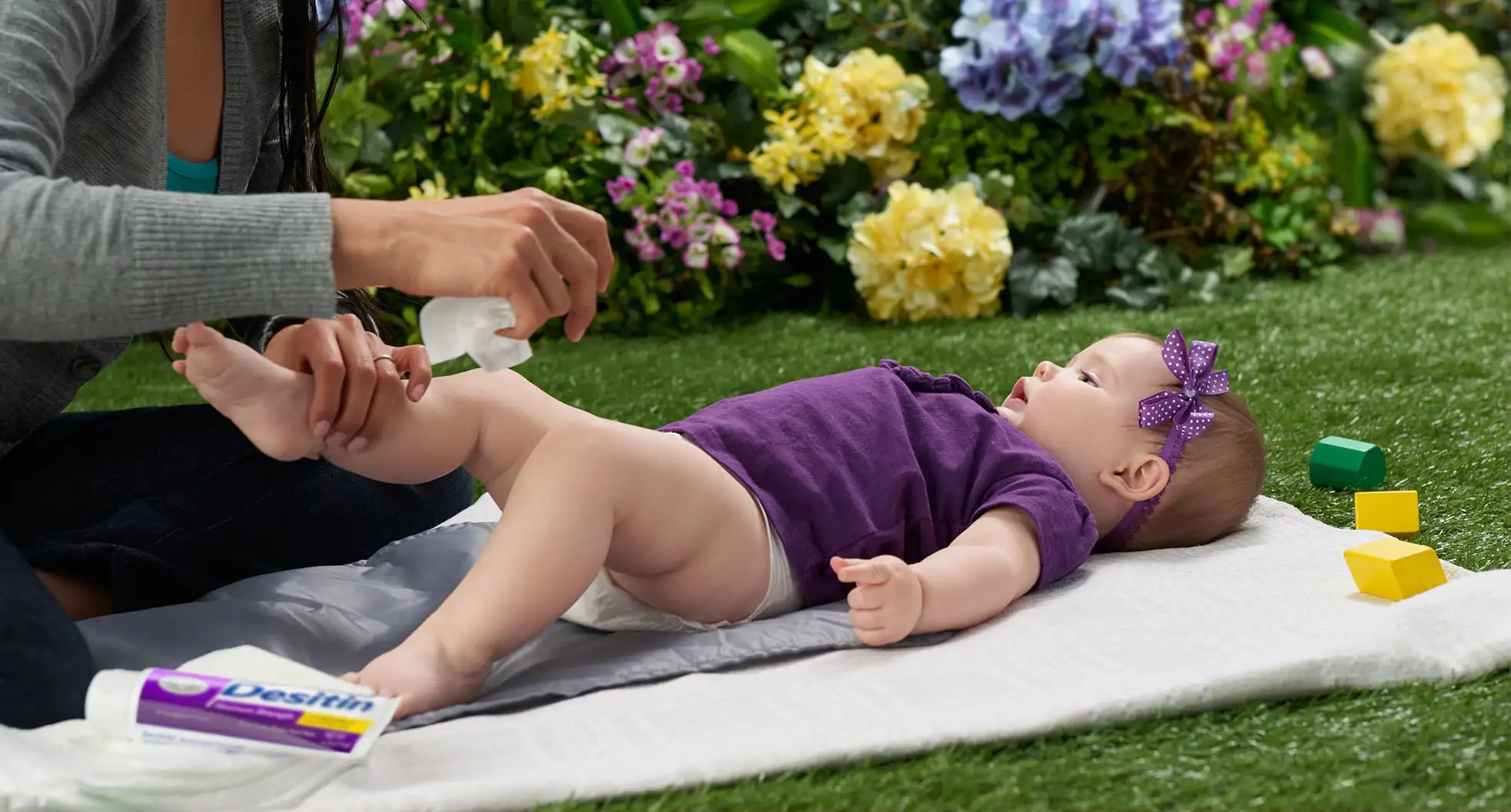 An adult changing a baby's diaper on a white mat in a garden with colorful flowers, DESITIN® Maximum Strength Original Zinc Oxide Paste and toy blocks around.