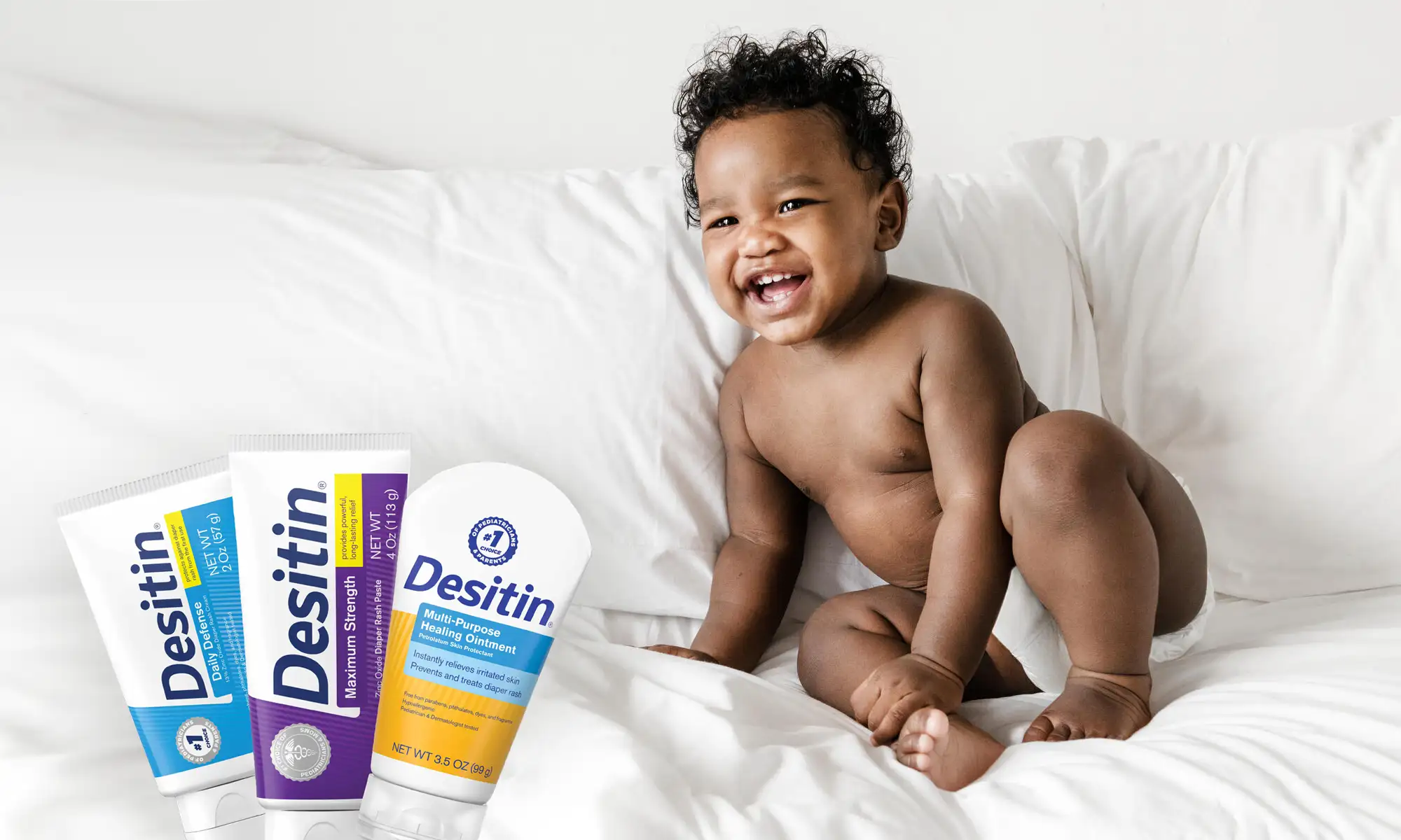 Desitin products are displayed beside a toddler sitting on a white bed, promoting skin care for babies. The packaging includes Maximum Strength and Multi-Purpose Healing Ointment tubes, emphasizing baby health and protection.