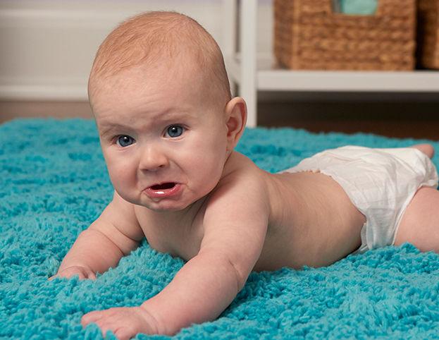 A baby in a diaper lying on a bright blue rug, looking up with a concerned or upset facial expression.