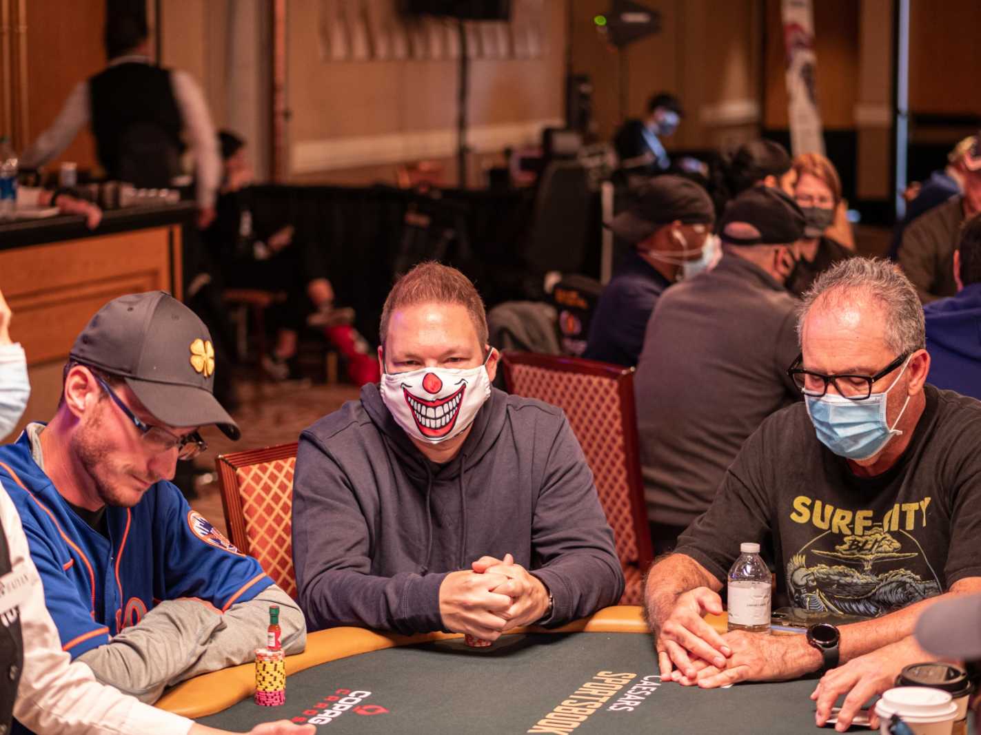 A poker player at the table wears a COVID mask bearing a clown's smile.