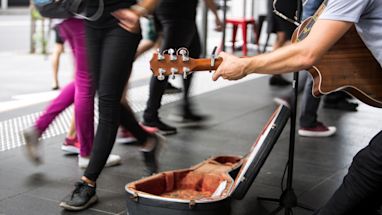 A street musician plays acoustic guitar next to an open guitar case as people walk by on a city pavement.