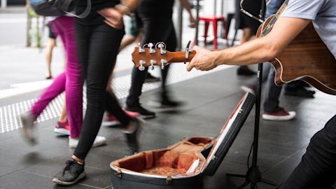 A street musician plays acoustic guitar next to an open guitar case as people walk by on a city pavement.
