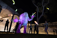 A large, illuminated purple puppet resembling a monster is manipulated by people at night, with jugglers tossing glowing objects nearby under an arched brick structure.