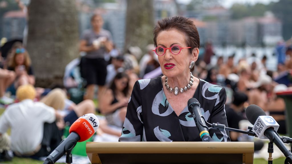 Clover Moore speaks at a podium with multiple microphones. A crowd is seated on grass in the background, near large trees and water.