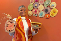 An Aboriginal Australian woman stands smiling, holding woven baskets, with colourful woven art pieces displayed on an orange wall behind her.