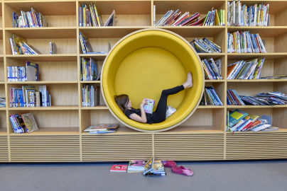 A person lounging inside a circular yellow reading nook, barefoot with a book, in a library. Books are neatly arranged on shelves around, with a few scattered on the floor.