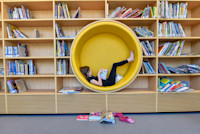 A person lounging inside a circular yellow reading nook, barefoot with a book, in a library. Books are neatly arranged on shelves around, with a few scattered on the floor.