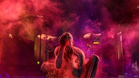 An Aboriginal dancer crouches low on stage over a cultural fire of smoking Eucalyptus leaves, cast in moody red and purple light
