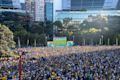 Large crowd gathers in an outdoor urban area to watch a soccer match displayed on a big screen, with modern buildings and trees in the background.