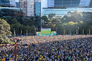 Large crowd gathers in an outdoor urban area to watch a soccer match displayed on a big screen, with modern buildings and trees in the background.