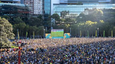Large crowd gathers in an outdoor urban area to watch a soccer match displayed on a big screen, with modern buildings and trees in the background.