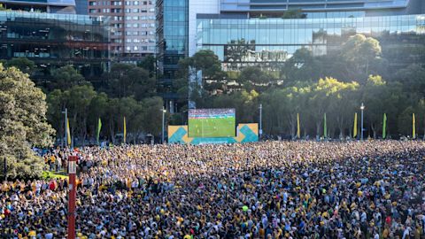 Large crowd gathers in an outdoor urban area to watch a soccer match displayed on a big screen, with modern buildings and trees in the background.
