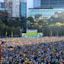 Large crowd gathers in an outdoor urban area to watch a soccer match displayed on a big screen, with modern buildings and trees in the background.