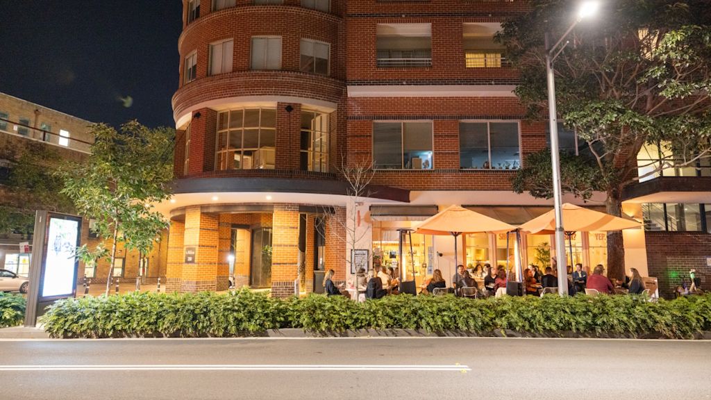 People dine outside under orange umbrellas at a restaurant on the ground floor of a curved brick building at night.